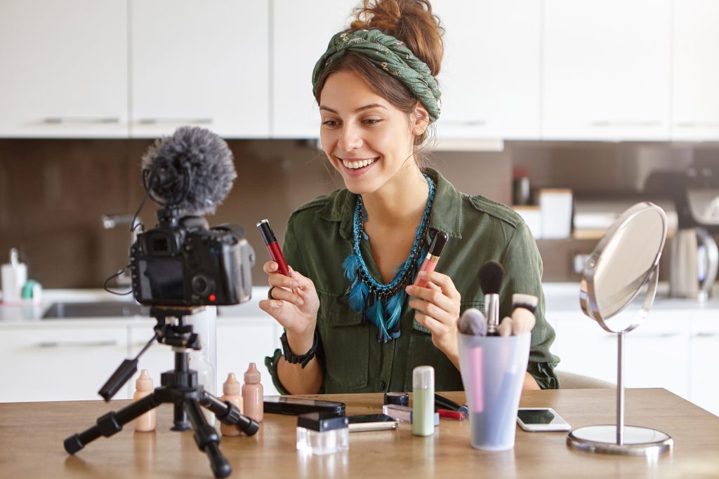 Young pretty female vlogger showing new kind of lipstick during recording vlog sitting at home. Woman shooting video indoors, advertising cosmetic products in front of camera, applying cosmetics
